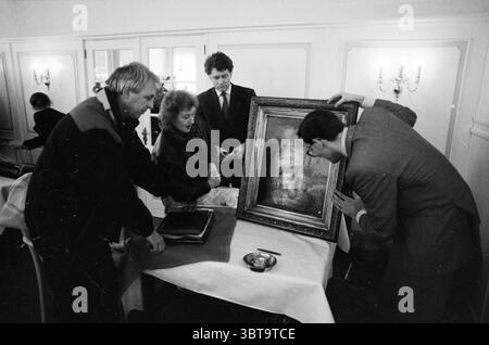Bewertungstag Christie's Groenendaal, Whizgle News, Dutch Desk, Niederlande, 1950 - 2000 am 13-02-1991. Das Bild enthält diese Themen. In einem warm beleuchteten Raum mit eleganter Einrichtung können vier Personen an einem Tisch aktiv werden. Die Umgebung lässt eine formelle Zusammenkunft vermuten, möglicherweise in einem privaten Speisesaal oder einer Kunstgalerie, die durch sanfte Beleuchtung von Wandlampen gekennzeichnet ist, die ein sanftes Leuchten auf die Szene wirft. An der Vorderseite sind zwei Männer und eine Frau um ein großes, reich verziertes Gemälde gedrängt, das gerade von seiner Schutzhülle entfernt wurde. Ein Mann, gekleidet in einem dunklen s Stockfoto