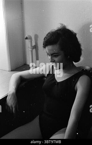 Interschool Konzerthalle Theater Education Theater, Whizgle News, Dutch Desk, Niederlande, 1950 - 2000 am 05-03-1963. Dies sind die Themen im Bild. In einem sanft beleuchteten Raum sitzt eine junge Frau bequem, ihre entspannte Haltung deutet auf ein Gefühl der Behaglichkeit und Besinnung hin. Sie trägt ein klassisches schwarzes Tanktop, das sich in einem Kontrast zu ihrer hellen Haut befindet, und ihr dunkles Haar umrahmt ihr Gesicht in weiche Locken. Das seitliche Profil zeigt ein sanftes Lächeln, das einen Ausdruck von Wärme und Zugänglichkeit vermittelt. Die Einrichtung besticht durch eine minimalistische Ästhetik mit hellen Wänden, die das geräumige Ambiente des verstärken Stockfoto