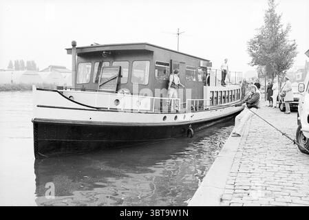 Living Barge Elisabeth auf dem Spaarne Kai Haarlem Spaarndamseweg Niederlande, Whizgle News, Dutch Desk, Niederlande, 1950 - 2000 am 19.09.1980. Das Bild enthält diese Themen. Die Szene zeigt eine ruhige Lage am Wasser, wo ein bescheidenes, klassisches Boot entlang eines Kopfsteinpflasterpfads anlegt, der vom ruhigen Wasser eines Kanals getrennt ist. Das Boot, hauptsächlich in einem tiefen Marineblau mit weißen Akzenten lackiert, verfügt über große Fenster mit Holzrahmen, was ihm einen malerischen Charme verleiht. Auf dem Boot sind ein paar Leute zu sehen. Einer steht aufmerksam am Rand und scheint eingeklemmt zu sein Stockfoto