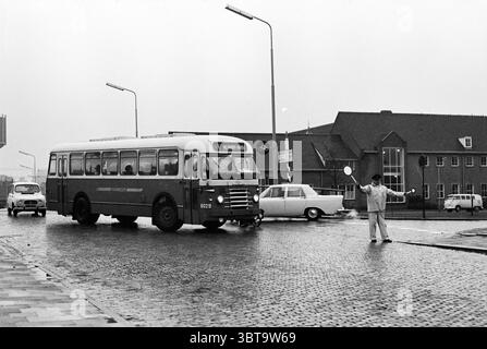 Bericht Naco Naco, Whizgle News, Dutch Desk, Niederlande, 1950 - 2000 am 11-11-1967. Dies sind die Elemente im Bild. Die Szene zeigt eine geschäftige Kreuzung an einem regnerischen Tag. Ein Oldtimer-Bus, der im Vordergrund deutlich zu sehen ist, hat ein markantes Rot-weiß-Farbschema. Die abgerundeten Kanten und das klassische Design erinnern an Nostalgie. Er ist an einer Kreuzung angehalten, was seine Rolle im öffentlichen Nahverkehr anzeigt. Links steht eine in leichter Kleidung gekleidete Person am Rande des Fußgängerübergangs und gibt Gesten, als ob sie den Verkehr lenkt oder Fußgängern assistiert. Ihre Haltung deutet auf eine se hin Stockfoto