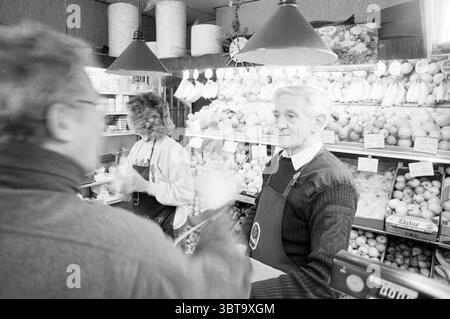 Interview mit Jaap Tanis Haarlem Smedestraat Niederlande, Whizgle News, Dutch Desk, Niederlande, 1950 - 2000 am 18.03.1989. Diese Themen werden in der Abbildung angezeigt. In einer belebten Marktszene führen zwei Personen eine lebhafte Interaktion an einem Obststand. Die Kulisse bietet eine lebhafte Präsentation frischer Produkte mit einer Auswahl an bunten Früchten wie Orangen, Bananen und Äpfeln, die ordentlich übereinander liegen. Die satten Farbtöne der Früchte stehen im Kontrast zu den gedämpften Tönen der Umgebung. Eine Person, ein älterer Gentleman, steht selbstbewusst hinter der Theke und trägt ein dunkles Schwitzen Stockfoto