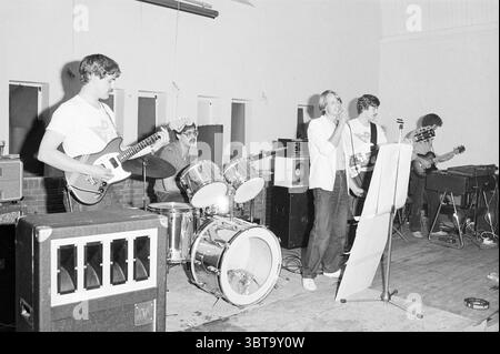 Performance 'Startseite' in Velsen-N' Music Velsen-Noord, Whizgle News, Dutch Desk, Niederlande, 1950 - 2000 am 29-05-1982. Dies sind die Themen im Bild. Die Szene zeigt eine lebhafte Band, die auf einer einfachen Bühne in einem Indoor-Veranstaltungsort mit einer rustikalen, entspannten Atmosphäre auftritt. Die Kulisse zeigt eine schlichte Wand und der Boden scheint aus Holz zu bestehen, was einen gemütlichen und einladenden Raum andeutet. Im Vordergrund steht ein männlicher Sänger in der Mitte, greift ein Mikrofon und engagiert sich aktiv mit der Menge. Er trägt ein lässiges Hemd und hat einen selbstbewussten Ausdruck, der Energie ausstrahlt. Bis Stockfoto