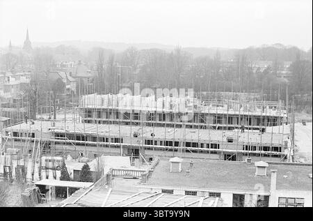 New Construction Houses Haarlem Nederland, Whizgle News, Dutch Desk, Niederlande, 1950 - 2000 am 19-03-1964. Diese Themen sind in der Abbildung dargestellt. Die Szene zeigt eine Baustelle voller Aktivität. Im Vordergrund ist ein Rahmen eines großen Bauwerks zu sehen, der mit Stahlträgern und Holzgerüsten deutlich aufsteigt und ein gitterähnliches Muster erzeugt. Die Skelettumrisse deuten auf die Anfänge eines mehrstöckigen Gebäudes hin, bei dem Arbeiter unter den Stützen verschiedene Aufgaben ausführen. Um das Gebäude herum befinden sich mehrere Erdhügel und Materialhaufen, darunter Dielen und Stockfoto