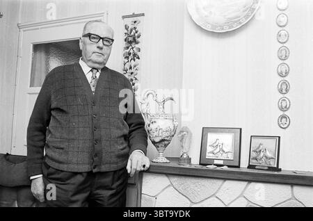Taubenfancier Van het Hof in Velsen Personen, Whizgle News, Dutch Desk, Niederlande, 1950 - 2000 am 10-12-1974. Diese Themen werden in der Abbildung angezeigt. Die Szene zeigt einen älteren Herrn, der selbstbewusst neben einem niedrigen Holzregal mit verschiedenen dekorativen Gegenständen steht. Seine Kleidung ist formell und doch lässig. Er hat eine dunkle, strukturierte Strickjacke über einem Hemd mit Kragen und eine gemusterte Krawatte. Die Brille des Mannes unterstreicht seinen nachdenklichen Ausdruck und verleiht ihm einen Hauch von Weisheit. Das Regal zeigt eine Vielzahl von Objekten, darunter eine große, verzierte Vase mit komplizierten Designs und zwei kleine fra Stockfoto