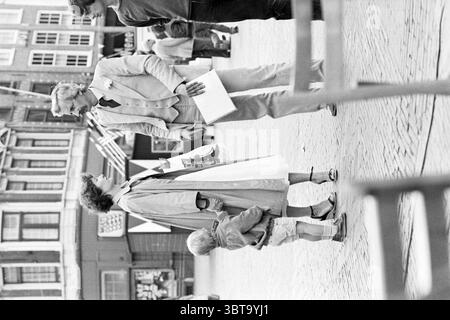 Haarlem Boekenstad (festlich gekleidete Familie) Haarlem Grote Markt The Netherlands, Whizgle News, Dutch Desk, Niederlande, 1950 - 2000 am 07-06-1979. Das Bild enthält diese Themen. In einer belebten Umgebung im Freien unterhalten sich zwei Erwachsene vor dem Hintergrund charmanter, alter Gebäude im klassischen Architekturstil. Die Szene strahlt ein Gefühl lebendiger sozialer Interaktion und Wärme aus. Die erste Figur, ein Mann in einem hellen, maßgeschneiderten Anzug, steht selbstbewusst. Er hat ein leichtes Lächeln und Gesten ausdrucksvoll, vielleicht macht er einen Punkt oder teilt eine fröhliche Anekdote. Sein Stockfoto
