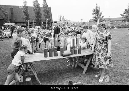 Spielplatz Oosterkwartier Ferienspiele Urlaubstage Ferienlager, Whizgle News, Dutch Desk, Niederlande, 1950 - 2000 am 20-07-1964. Das Bild enthält diese Themen. Die Szene fängt eine lebhafte Atmosphäre im Freien ein, mit einem großen, grasbewachsenen Bereich voller Aktivität. An der Vorderseite befindet sich ein langer Tisch aus Holz, der mehrere Holzzylinder in einer prekären Weise stapelt, was auf ein Spiel oder eine Herausforderung hinweist, bei dem Balance und Geschick gefragt sind. Um den Tisch herum versammelt sich eine Gruppe von Kindern, deren Ausdruck eine Mischung aus Spannung und Konzentration ist und die Aktivitäten eifrig beobachten Stockfoto