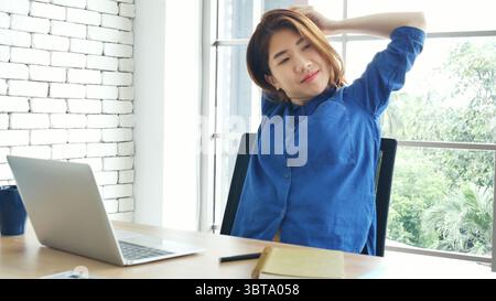 Frau streckt den Arm hoch und sitzt falsch in der Position des Home Office-Schreibtisches. Rückseite einer jungen asiatischen Frau müde von der Arbeit Körper Stress Rücken Schmerz Büro Sy Stockfoto