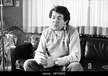 Portrait Professor van Dongen Santpoort Portraits Santpoort, Whizgle News, Dutch Desk, Niederlande, 1950 - 2000 am 08-12-1979. Diese Themen werden in der Abbildung angezeigt. Ein Mann sitzt bequem auf einer weichen schwarzen Couch, seine Haltung entspannt, aber nachdenklich. Er hat lockige, dunkle Haare und trägt einen hellgrauen Pullover über einem Hemd mit Kragen, was ihm ein lässiges, aber zusammengesetztes Aussehen verleiht. Seine Augen sind geschlossen, was auf einen Moment der Besinnung oder der Introspektion hindeutet. Um ihn herum gibt es weiche, neutrale Möbel, die eine warme und einladende Atmosphäre schaffen. Die Couch ist mit mehreren dekorativen Plättchen verziert Stockfoto