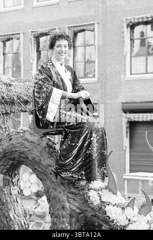 Tulip Queen auf KLM Parade Float Flower Parade Blumenausstellung, Whizgle News, Dutch Desk, Niederlande, 1950 - 2000 am 22-04-1988. Das Bild enthält diese Themen. In einer lebhaften Feier sitzt eine junge Frau auf einem großen Wagen und verkörpert einen Hauch von Eleganz. Sie trägt ein fließendes Kleid, das mit komplizierten Mustern verziert ist und einen reichen Kontrast zu den gedämpften Tönen ihrer Umgebung zeigt. Der Stoff blitzt und fängt das Licht auf eine Weise ein, die seine Schönheit unterstreicht. Der Schwimmer selbst ist mit einer Vielzahl von bunten Blumen dekoriert, die sich durch das Design mit organischen Kurven winden und die Verschönerung verstärken Stockfoto