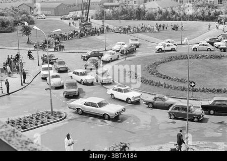 Verkehrsstau an einem Kreisverkehr. Whizgle News, Dutch Desk, Niederlande, 1950 - 2000. Das Bild enthält diese Themen. In dieser belebten Szene ist eine kreisförmige Kreuzung mit einer Vielzahl von Fahrzeugen und einer Menschenmenge gefüllt. Die Komposition zeigt eine Mischung aus Oldtimern aus verschiedenen Epochen mit verschiedenen Formen und Farben – einige in weichen Pastelltönen, während andere eher dezent oder lebhaft sind. Ihre glänzenden Oberflächen und eleganten Linien reflektieren das Licht und fügen der Szene ein dynamisches Element hinzu. Rund um den Kreisverkehr bilden gepflegtes Grün und ordentlich angeordnete Blumenbeete wieder einen Kontrast Stockfoto