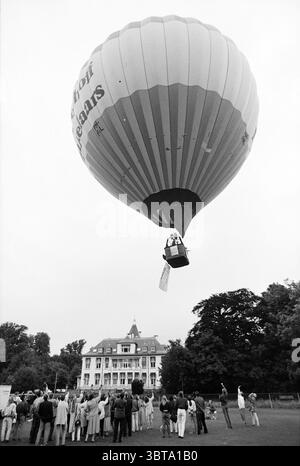 Direktflug im Ballon[eur] Beijer, Whizgle News, Dutch Desk, Niederlande, 1950 - 2000 am 24-06-1993. Dies sind die Themen im Bild. In der Szene dominiert ein lebhafter Heißluftballon den Himmel, dessen große, runde Form mit bunten Streifen gefüllt ist, die von warmen Rottönen bis zu kühlen Blautönen reichen. Der Ballon ist mit einem Fesseln verbunden und schwebt direkt über einem üppig grünen Rasen, wo sich eine vielfältige Gruppe von Schaulustigen unter sich sammelt. Das Publikum, bestehend aus Männern und Frauen verschiedener Altersgruppen, ist in einer Mischung aus lässiger und leicht formeller Kleidung gekleidet. Einige Menschen zeigen nach oben, ihre Gesichter zeigen einen Ausdruck der Erregung Stockfoto