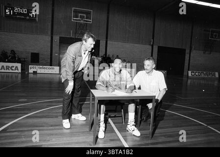 Vertrag mit Basketball Club, Whizgle News, Dutch Desk, Niederlande, 1950 - 2000 am 27-04-1990. Dies sind die Themen im Bild. Die Szene fängt einen dynamischen Moment in einem Fitnessraum ein, der durch einen Holzboden gekennzeichnet ist, der die Umgebungsbeleuchtung reflektiert. In der Mitte sitzt ein junger Mann an einem einfachen Tisch, lässig gekleidet in einem sportlichen Outfit, wahrscheinlich ein Trikot, kombiniert mit Shorts und Sportschuhen. Sein Ausdruck ist fokussiert und entschlossen, was auf die Bedeutung des Moments hindeutet, den er erlebt. Er wird von zwei Männern begleitet, die beide mit ihm reden. Eins, links, A Stockfoto