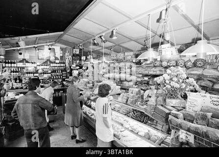 Int. Bäckerei, Whizgle News, Dutch Desk, Niederlande, 1950 - 2000 im Jahr 1976. Dies sind die Themen im Bild. Die Szene fängt einen belebten Indoor-Markt ein, wahrscheinlich ein Delikatessen- oder Lebensmittelladen, der durch eine lebendige Ausstellung von Lebensmitteln gekennzeichnet ist. Im vorderen Teil des Glaskastens finden Sie eine große Auswahl an Käse und Aufschnitt, die in verlockenden Blöcken und Scheiben angeordnet sind. Die Auswahl ist vielfältig, mit Texturen von glatt und cremig bis fest und bröckig, alle in Gelb-, weiß- und Brauntönen, durchsetzt mit Akzenten von Kräutern und Gewürzen. Hinter dem Tresen ist ein Anbieter Acti Stockfoto
