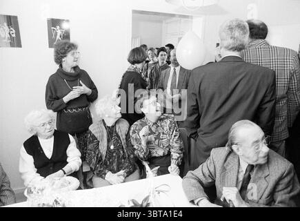 Offenes Wohnzimmer, Whizgle News, Dutch Desk, Niederlande, 1950 - 2000 am 11-02-1994. Das Bild enthält diese Themen. Die Szene fängt ein lebhaftes Treffen in einem hellen, luftigen Raum ein. Die Wände sind mit einfachen Dekorationen geschmückt, und die Atmosphäre ist erfüllt von einem Gefühl der Feier und Wärme. Im Vordergrund sitzt eine Gruppe älterer Personen um einen Tisch, deren Ausdrucksformen animiert und engagiert sind. Eine Frau, die eine Brille und eine dunkle Weste über einer hellen Bluse trägt, scheint eine Geschichte zu teilen, wobei ihre Hände ausdrucksvoll wirken. Neben ihr, eine andere Frau, mit grauem Haar elegant gestylt Stockfoto