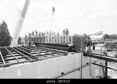 Neubau Zuckerfabrik Halfweg C.M.S. Halfway, Whizgle News, Dutch Desk, Niederlande, 1950 - 2000 am 05-09-1978. Das Bild enthält diese Themen. Die Szene zeigt eine industrielle Bauumgebung unter hellem Tageslicht. Ein großer Kran dominiert den oberen Teil der Komposition, dessen Arm sich nach außen erstreckt und eine massive Metallkonstruktion trägt, die in Position gebracht wird. Das Metall wirkt dunkel und glänzend und kontrastiert mit den helleren Tönen der Umgebung. Auf der linken Seite sind zwei Arbeiter sichtbar, die in Sicherheitsausrüstung gekleidet sind, einschließlich Schutzhüten und Kleidung, die auf einen hindeutet Stockfoto