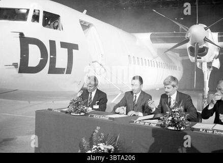 Vertrag Züchter DLT Fokker, Whizgle News, Dutch Desk, Niederlande, 1950 - 2000 am 07-08-1987. Dies sind die Elemente im Bild. Die Szene fängt einen Moment einer wichtigen Ankündigung oder eines Ereignisses ein, wahrscheinlich im Kontext der Luftfahrt. Ein großes Flugzeug, das sich durch seinen schlanken Körper und markante Propeller auszeichnet, nimmt den Hintergrund ein. Die sichtbaren Buchstaben „DLT“ sind kühn an der Seite des Flugzeugs angebracht und deuten auf eine Unternehmens- oder Fluggesellschaft hin. Im Vordergrund sitzen drei Personen an einem langen Tisch, der mit kleinen Blumenarrangements geschmückt ist, die einen Hauch von Farbe verleihen Stockfoto