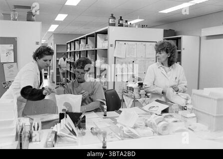 Direktor des Pharmazie-Instituts der Haarlem Krankenhäuser Haarlem Niederlande, Whizgle News, Dutch Desk, Niederlande, 1950 - 2000 am 20-11-1996. Dies sind die Elemente im Bild. Die Szene erfasst eine lebendige Arbeitsumgebung in einer medizinischen oder Laborumgebung. Drei Personen sind an einem überdachten zentralen Tisch, umgeben von Regalen, die mit medizinischen Vorräten und organisiertem Material gefüllt sind, mit verschiedenen Aufgaben beschäftigt. Die Individuen zeigen unterschiedliche Ausdrucksformen von Konzentration und Konzentration. Auf der linken Seite lehnt sich eine Person in einem weißen Labormantel nach vorne und beobachtet aufmerksam die Arbeit der nächsten Person Stockfoto