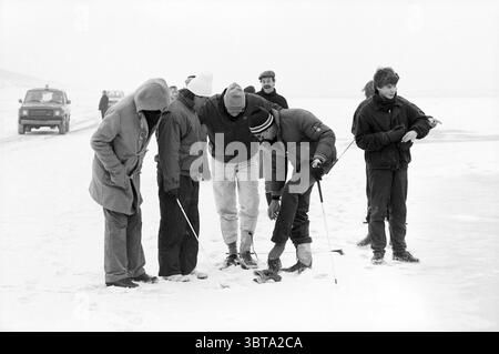 Golf am Noordwijk Beach - Zandvoort Golf Golf Golf Noordwijk, Whizgle News, Dutch Desk, Niederlande, 1950 - 2000 am 22-02-1986. Das Bild enthält diese Themen. In einer kahlen, winterlichen Umgebung versammelt sich eine Gruppe von sechs Individuen auf einer gefrorenen Eisfläche. Die Szene ist von einer gedämpften, fast monochromatischen Palette von weiß- und Grautönen umgeben, die auf eine schwere Schneedecke hindeuten. Die Menschen sind in verschiedenen Winterbekleidung gebündelt, dicke Mäntel und Kapuzen gegen die Kälte gezogen. Einige tragen Mützen oder Hüte fest nach unten gezogen, was das Gefühl der Kälte in der Luft erhöht. Ihre Körperhaltungen Stockfoto