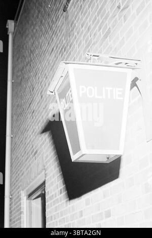 Außenlampe eines Polizeipostens Heemskerk, Whizgle News, Dutch Desk, Niederlande, 1950 - 2000 am 15-10-1968. Dies sind die Elemente im Bild. Die Szene zeigt eine Wand aus strukturiertem, rotbraunem Ziegelstein, die eine raue Kulisse schafft, die eine Aura von Beständigkeit und Stabilität ausstrahlt. Ein deutliches, beleuchtetes Schild hängt von der Wand, nach außen geneigt. Seine geometrische Struktur erinnert an eine Laterne mit einem rechteckigen Körper, der sich leicht verjüngt. Die Hauptfarbe des Schildes ist ein helles, markantes Rot, das sich in einem scharfen Kontrast zu den dunkleren Tönen der Ziegelsteine und der Umrandung bildet Stockfoto