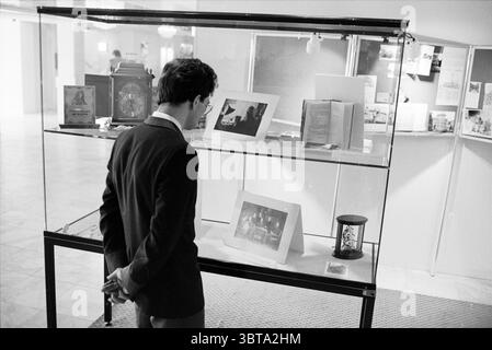 Ausstellung von Corrie Ten Boom im Hotel Barbizon Hoofddorp Museum Museen Hoofddorp Niederlande, Whizgle News, Dutch Desk, Niederlande, 1950 - 2000 am 06-02-1988. Diese Themen werden in der Abbildung angezeigt. Die Szene zeigt einen Mann, der in einem dunklen Anzug gekleidet ist und einen Blick auf eine Vitrine voller Artefakte und gerahmter Bilder wirft. Die Beleuchtung ist weich und gestreut, was eine ruhige Atmosphäre im Raum schafft. Der Mann steht mit dem Rücken zum Betrachter, seine Hände sind hinter ihm gefesselt, was ein Gefühl von Neugier und Nachsicht andeutet. Seine Frisur ist ordentlich, und er scheint sich auf den konzentriert zu haben Stockfoto