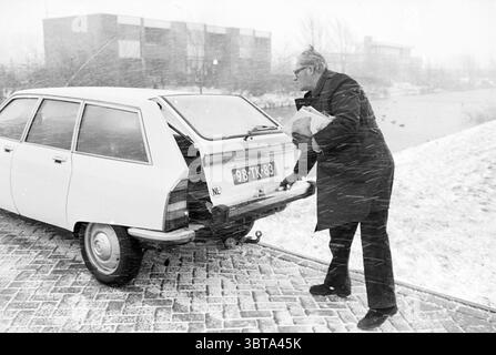 VNU Magazin Lieferperson im Schnee. VNU Association of Dutch Publishers, Whizgle News, Dutch Desk, Niederlande, 1950 - 2000 am 10-01-1979. Die Abbildung zeigt diese Themen. Ein Mann, in dunkler Kleidung gekleidet, steht neben einem weißen Fließheck-Auto, das teilweise auf einem Kopfsteinpflasterweg geparkt ist. Er scheint ein Objekt im Fond des Fahrzeugs zu finden oder zu platzieren. Die Szene spielt inmitten eines starken Schneefalls mit großen, flauschigen Flocken, die schnell fallen, den Boden bedecken und die Oberflächen in einer dicken weißen Schicht verdecken. Die Umgebung ist mit Grautönen gedämpft Stockfoto