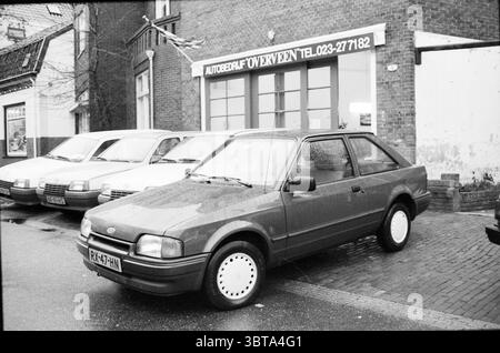 Autobedrijf Overveen mit Autos vor der Tür Overveen, Whizgle News, Dutch Desk, Niederlande, 1950 - 2000 am 20-12-1989. Die Abbildung zeigt diese Themen. In einer belebten städtischen Umgebung steht ein kompaktes Auto im Vordergrund. Seine schlanken, abgerundeten Konturen werden durch eine tiefe, satte Farbe, vielleicht einen Rot- oder Kastanienton, hervorgehoben, die sich von den gedämpften Tönen der Umgebung abhebt. Das Fahrzeug steht auf einer leicht feuchten Asphaltoberfläche, was auf jüngste Regenfälle hindeutet, wobei Regentropfen auf der Oberfläche glitzern. Hinter dem Wagen befinden sich mehrere andere Fahrzeuge Stockfoto