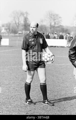 Football Kennemerland-DCS, Whizgle News, Dutch Desk, Niederlande, 1950 - 2000 am 21-03-1997. Dies sind die Themen im Bild. Die Szene zeigt einen Mann in einer dunklen Streifenuniform, der auf einem grasbewachsenen Sportplatz steht. Er hält einen Fußball unter einem Arm, sein Ausdruck ernst und konzentriert. Die Umgebung ist ein klarer Tag, bei dem sanftes Sonnenlicht die Szene beleuchtet und sanfte Schatten auf den Boden wirft. Im Hintergrund sind unscharfe Zahlen von Menschen zu sehen, wahrscheinlich von Zuschauern oder Teammitgliedern, die ein Gefühl der Gemeinschaft und gemeinsame Vorfreude schaffen. Die Farben sind hauptsächlich stummgeschaltet, mit Stockfoto