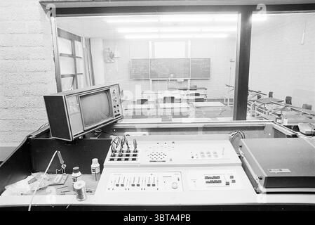 Innenpädagogische Akademie 'de la Salle' Interior, Whizgle News, Dutch Desk, Niederlande, 1950 - 2000 am 18-01-1977. Das Bild enthält diese Themen. Die Einrichtung ist ein Kontrollraum mit Blick auf einen Klassenzimmer. Im Vordergrund befindet sich eine dunkle, flache Oberfläche, die verschiedene Geräte enthält, darunter ein Vintage-Fernsehgerät, das eine gedämpfte Szene zeigt. Der Fernseher hat ein Kastendesign und ein analoges Erscheinungsbild, was auf eine frühere Ära hinweist. Daneben befindet sich ein Bedienfeld mit einer Reihe von Tasten, Knöpfen und Schiebereglern, die ein Gefühl von Funktionalität und Komplexität vermitteln. Der Hintergrund zeigt sich Stockfoto