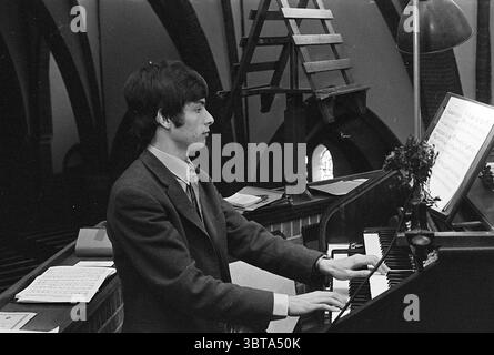 Organist Frans Geels Orgeln und Organisten, Whizgle News, Dutch Desk, Niederlande, 1950 - 2000 am 01-06-1970. Dies sind die Elemente im Bild. In einem schwach beleuchteten Raum, der einen Charme der alten Welt ausstrahlt, konzentriert sich ein junger Mann intensiv auf das Klavierspielen. Er hat dunkle, zerzauste Haare und trägt einen maßgeschneiderten Anzug, der auf einen formellen Anlass hinweist. Das Ambiente ist warm und doch gedämpft, beleuchtet durch sanfte Deckenbeleuchtung, die sanfte Schatten wirft. Zu seiner Rechten liegt eine Reihe von Noten verstreut, wobei einige Blätter gegeneinander angesprungen sind, während im Hintergrund ein teilweise ausgedehnter Musikstand steht Stockfoto