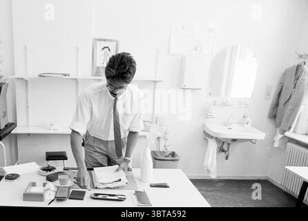 Militärkrankenhaus Overveen|Marine Krankenhaus Overveen Overveen Bloemendaalseweg, Whizgle News, Dutch Desk, Niederlande, 1950 - 2000 am 23-05-1990. Das Bild enthält diese Themen. In einem hellen und minimalistischen Raum steht ein Mann an einem weißen Tisch mit verstreuten Papieren und verschiedenen Gegenständen. Er hat kurze, ordentlich gestylte Haare und trägt eine Brille in einem knusprigen weißen Hemd, das sich von seiner dunkleren Hose abhebt. Die Atmosphäre fühlt sich ruhig und doch beschäftigt an, da er sich intensiv darauf konzentriert, die Dokumente vor ihm zu sortieren. Die Wände sind mit ein paar einfachen Zeichnungen und Notizen geschmückt Stockfoto
