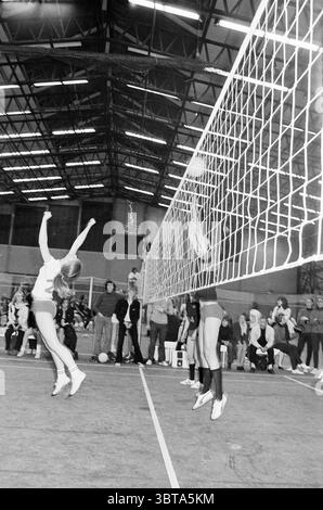 Volley-Ball., Whizgle News, Dutch Desk, Niederlande, 1950 - 2000. Diese Themen sind in der Abbildung dargestellt. Die Szene fängt einen dynamischen Moment in einem Indoor-Volleyballspiel ein, das durch die hohe Energie und die Athletik der Spieler gekennzeichnet ist. Im Vordergrund werden zwei Athleten hervorgehoben: Ein Spieler springt in die Luft, die Arme sind in Richtung Volleyballnetz gestreckt, während der andere den Schuss blockiert. Beide tragen sportliche Kleidung, wobei eines in einem hellen Oberteil und Shorts, das andere in dunkleren Shorts und einem taillierten Oberteil einen Kontrast zum Hintergrund bildet. Die Spieler strahlen aus Stockfoto