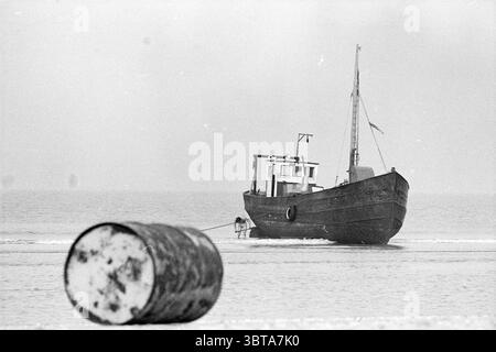 Stranded Ship Parnassia Beach Ships, Whizgle News, Dutch Desk, Niederlande, 1950 - 2000 am 16-10-1974. Dies sind die Elemente im Bild. Die Szene zeigt eine ruhige Küstenlandschaft, in der ein kleines Boot sanft am Ufer des Wassers liegt. Das Boot ist in dunklen Tönen lackiert und hat ein verwittertes Aussehen, was darauf hindeutet, dass es im Laufe der Zeit viele Gewässer durchquert hat. Seine Silhouette hebt sich vor einem trüben Hintergrund ab und fügt sich subtil in die gedämpften Farben der Szene ein. Im Vordergrund liegt ein großes Fass teilweise in den Sand getaucht. Der Zylinder ist durch die Zeit gekennzeichnet, seine Oberfläche weist Rostflecken und auf Stockfoto