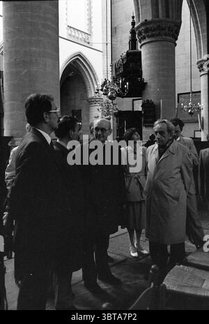 Bischof Bomers besucht St. Bavo Churches Roman Catholic Church, Whizgle News, Dutch Desk, Niederlande, 1950 - 2000 am 20.05.1984. Das Bild enthält diese Themen. Die Szene entfaltet sich in einem geräumigen, gewölbten Innenraum, das ein Gefühl von Geschichte und Hochmut ausstrahlt, geprägt von Steinbögen und hohen Decken. Natürliches Licht filtert durch große, komplizierte Fenster und erzeugt sanfte Muster auf den Steinboden, die der ansonsten kühlen Umgebung Wärme verleihen. Im Vordergrund steht eine kleine Gruppe von Personen im Gespräch. Sie tragen eine Mischung aus formeller und halbformaler Kleidung, Stockfoto