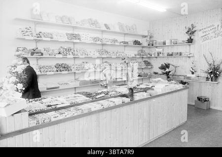 Interior Candy King IJmuiden Interior, Whizgle News, Dutch Desk, Niederlande, 1950 - 2000 am 04-03-1970. Das Bild enthält diese Themen. Die Szene spielt in einem malerischen Süßwarengeschäft, das von einer warmen, nostalgischen Atmosphäre geprägt ist. Die Wände sind mit hellen Holztäfelungen gesäumt, die eine natürliche und einladende Atmosphäre verleihen. Die Regale erstrecken sich über den gesamten Raum, gefüllt mit einer bunten Auswahl an Süßigkeiten, Süßigkeiten und verpackten Leckereien, die ein visuelles fest für die Augen schaffen. Das Display zeigt eine Mischung aus Texturen und Formen; Gläser mit bunten Gummigummis, Schokoladentafeln in glänzender Folie, Stockfoto