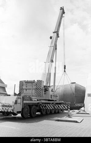 Große Transportunternehmen IJmuiden Hoogovens 6 Transportunternehmen IJmuiden the Netherlands, Whizgle News, Dutch Desk, Niederlande, 1950 - 2000 am 10-07-1985. Die Abbildung zeigt diese Themen. Die Szene wird von einem großen Radkran dominiert, der auf einer gepflasterten Fläche positioniert ist. Die hoch aufragende Struktur des Krans erstreckt sich hoch in den Himmel und verfügt über einen langen ausziehbaren Arm, der sich zu einem großen, dunklen, kugelförmigen Objekt ausbreitet, das angehoben wird. Das Objekt wirkt robust und industriell und zeichnet sich durch eine glatte, gekrümmte Oberfläche aus, die das umgebende Licht subtil reflektiert. Um den Kran und das Lift herum Stockfoto