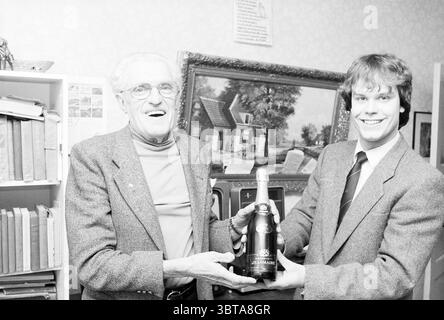 Eine Flasche Champagner für Mr., Whizgle News, Dutch Desk, Niederlande, 1950 - 2000 am 12-1986. Diese Themen sind in der Abbildung dargestellt. In einem warm beleuchteten Innenraum werden zwei Personen in der Mitte der Szene sichtbar gezeigt, die einen Moment der Feier verbringen. Der ältere Herr mit silbernen Haaren und Brillen lächelt lebhaft und strahlt ein Gefühl von Freude und Wärme aus. Er trägt ein helles Button-up-Shirt unter einem strukturierten Blazer, das sich von den dunkleren Tönen um ihn herum abhebt. Neben ihm steht ein jüngerer Mann, gepflegt mit freundlichem Auftreten, gekleidet in einen Anzug mit Stockfoto