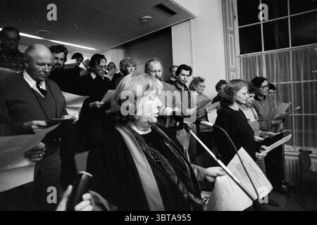 Singen Sie Protest von Nachbarschaftsräten im Rathaus mit Bürgermeister Schmitz Haarlem the Netherlands, Whizgle News, Dutch Desk, Niederlande, 1950 - 2000 am 24.11.1991. Dies sind die Elemente im Bild. Die Szene fängt eine Gruppe von Individuen ein, die sich in einem etwas schwach beleuchteten Raum versammelt haben, mit einem Fokus auf eine Frau, die an der Vorderseite steht. Sie befindet sich etwas rechts von der Mitte, hält ein Mikrofon und blickt auf das Publikum hin und strahlt ein Gefühl von Autorität und Präsenz aus. Die Frau scheint die Gruppe zu führen, die aus Männern und Frauen unterschiedlichen Alters besteht, von denen viele halten Stockfoto
