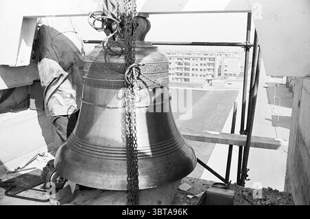 Römisch-katholische Kirche Beverwijk Baustellen römisch-katholische Kirchen römisch-katholische Kirchen Uhren und Uhren, Whizgle News, Dutch Desk, Niederlande, 1950 - 2000 am 09-04-1964. Das Bild enthält diese Themen. Die Szene nimmt eine große Glocke auf, die deutlich sichtbar dargestellt wird, während sie an einer Stützstruktur hängt. Diese Glocke hat eine polierte, metallische Oberfläche, die das Umgebungslicht reflektiert und trotz der monochromatischen Töne einen warmen Glanz erzeugt. Um die Glocke herum bilden Holzbalken und Metallgerüste einen Rahmen, der die industriellen Aspekte der Installation unterstreicht Stockfoto