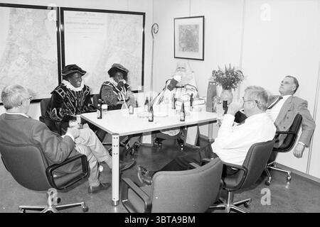 Sint HD, Whizgle News, Dutch Desk, Niederlande, 1950 - 2000 am 03-02-1991. Diese Themen werden in der Abbildung angezeigt. In einem geräumigen, gut beleuchteten Raum nimmt eine Gruppe von sechs Personen einen zentralen Tisch ein, der mit mehreren leeren Flaschen ordentlich angeordnet ist. Die Atmosphäre scheint entspannt zu sein, alle unterhalten sich, einige halten Getränke. Die Menschen am Tisch zeigen eine Vielzahl von Stilen und Ausdrücken. Auf der einen Seite sind drei Individuen in auffälligen Kostümen mit satten, dunklen Tönen und farbenfrohen Ornamenten verziert. Ihre Kleidung umfasst ausgeklügelte Hüte, die einen fe hinzufügen Stockfoto