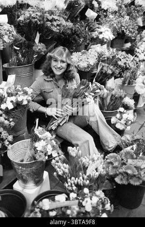 Junge Frau unter Blumen vor einem Geschäft ausgestellt, Whizgle News, Dutch Desk, Niederlande, 1950 - 2000 am 19-03-1979. Das Bild enthält diese Themen. In einem lebhaften Blumenladen ist die Szene mit einer Vielzahl von leuchtenden Blumen in verschiedenen Farbtönen gefüllt, die einen farbenfrohen Wandteppich der Natur schaffen. Die Umgebung ist geschäftig, mit zahlreichen Eimern und Töpfen voller Blüten – Rosen, Tulpen und Gänseblüten in Rosa-, Gelb- und Weißtönen. In der Mitte sitzt eine Frau bequem auf dem Boden, umgeben von diesen Blumenarrangements. Sie hat helles, welliges Haar und ist in einer lässigen, hellen Farbe gekleidet Stockfoto