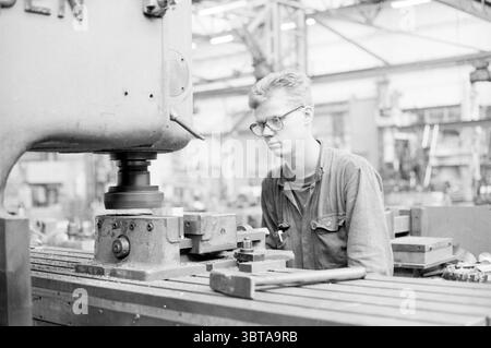 Maschinenbau Stahlarbeiter Figee, Whizgle News, Dutch Desk, Niederlande, 1950 - 2000 am 05-09-1991. Das Bild enthält diese Themen. In einer geschäftigen industriellen Umgebung steht ein junger Mann auf eine schwere Maschine konzentriert, die tief in seine Arbeit verwickelt ist. Er trägt funktionelle Kleidung, die sich durch ein festes Hemd mit Knöpfen, Ärmel bis zu den Ellbogen gerollt und eine Schutzbrille auf der Nase auszeichnet, was seine Konzentration unterstreicht. Die Maschine vor ihm ist in ihrer Größe von großer Bedeutung und verfügt über eine graue Metallic-Oberfläche, die das Umgebungslicht reflektiert. Es besteht aus mehreren großen mechanischen Komponenten, darunter einem Hea Stockfoto