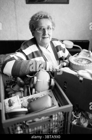 Frau van Empel, Whizgle News, Dutch Desk, Niederlande, 1950 - 2000 am 02-03-1992. Diese Themen werden in der Abbildung angezeigt. In einem gemütlichen Innenraum sitzt eine Seniorin bequem inmitten einer Sammlung verschiedener Objekte. Sie hat weiche, lockige graue Haare und trägt eine große, runde Brille, die ihr warmes Lächeln unterstreicht. Ihre Kleidung ist mit einer gestreiften Strickjacke ausgestattet, die überwiegend in hellen und dunklen Tönen gehalten ist, was ihrem Aussehen eine Dimension der Textur verleiht. Um sie herum sind zwei Körbe gefüllt mit einem eklektischen Sortiment. Die Körbe, einer in dunkelgrauem und der andere in hellerem Schatten, überlaufen mit Gegenständen, die darin enthalten sind Stockfoto