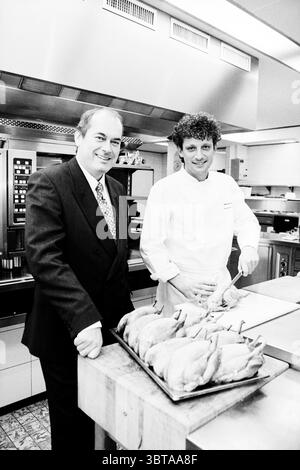 J. Beeren + Rive Kok, Whizgle News, Dutch Desk, Niederlande, 1950 - 2000 am 03-12-1997. Dies sind die Elemente im Bild. In einer hellen Küche stehen zwei Personen an einer Holztheke. Die Atmosphäre ist lebhaft, gefüllt mit den Düften des Essens und den Klängen der kulinarischen Aktivität. Eine Person, die in einer weißen Küchenuniform gekleidet ist, scheint konzentriert und engagiert zu sein und geht geschickt mit einem ganzen Huhn um. Der Koch hat lockiges Haar und zeigt einen konzentrierten Blick, was auf Fachwissen in seiner Handwerkskunst hindeutet. Neben ihm steht ein Mann in einem gut geschnittenen dunklen Anzug, der Selbstvertrauen ausstrahlt, mit einem Tuch Stockfoto