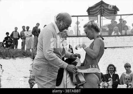 Miss Beach Election Zandvoort II Zandvoort Zandvoort, Whizgle News, Dutch Desk, Niederlande, 1950 - 2000 am 08-08-1981. Das Bild enthält diese Themen. In der Szene steht eine Frau im Badeanzug im Mittelpunkt der Aufmerksamkeit, die einen Preis oder eine Auszeichnung von einem angesehenen älteren Gentleman erhält. Sie hat ein kurzes, helles Haar und zeigt eine Pose, die eine Mischung aus Stolz und vielleicht leichter Schüchternheit zeigt. Der Gentleman, gekleidet in einem hellen Pullover und einer Brille, hat einen warmen Ausdruck, wenn er ihr ein Objekt übergibt, wahrscheinlich eine Schärpe oder ein Band. Um sie herum versammelt sich eine vielfältige Gruppe von Zuschauern, s Stockfoto