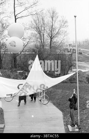 Eröffnung des Radweges Schiphol Schiphol, Whizgle News, Dutch Desk, Niederlande, 1950 - 2000 am 25-04-1994. Dies sind die Elemente im Bild. Die Szene fängt einen düsteren und bewölkten Tag ein, der durch eine gedämpfte Farbpalette aus Grau- und Brauntönen gekennzeichnet ist. Eine Gruppe von Individuen versammelt sich um einen großen weißen Stoff, der von einigen Leuten aufgehalten wird. Der Stoff scheint etwas Feuchtigkeit angesammelt zu haben, was zu der trüben Atmosphäre beiträgt, und seine Oberfläche ist leicht zerknittert, was darauf hindeutet, dass er vor kurzem aufgerollt oder im Wind gefangen wurde. Eine Person, in dunkler Kleidung gekleidet, steht an erster Stelle ich Stockfoto
