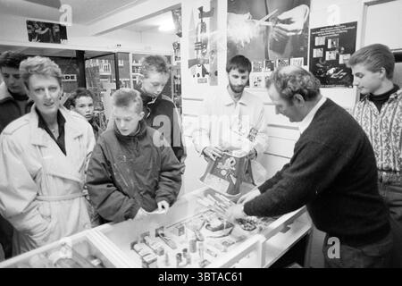 Feuerwerk Verkauf Jupiter Zandvoort Feuerwerk Zandvoort, Whizgle News, Dutch Desk, Niederlande, 1950 - 2000 am 29-12-1989. Dies sind die Elemente im Bild. Die Szene fängt eine geschäftige Innenumgebung ein, wahrscheinlich ein kleines Geschäft oder Geschäft mit Schwerpunkt auf persönlichen Interaktionen. Eine Gruppe von Personen besetzt den Vordergrund, die sich um eine Vitrine mit bunten Gegenständen versammelt haben. Die Vitrine ist transparent und ermöglicht die Sichtbarkeit verschiedener Produkte, darunter Snacks oder Schmuckstücke, und schafft so eine einladende und zugleich energiegeladene Atmosphäre. In der Mitte scheint ein Mann in einem schwarzen Pullover verwickelt zu sein Stockfoto