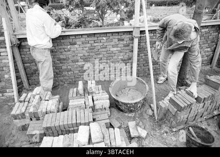Renovierung der Gartenwand Oud Velsen Velsen, Whizgle News, Dutch Desk, Niederlande, 1950 - 2000 am 27-05-1991. Diese Themen werden in der Abbildung angezeigt. In einer belebten Bauszene konzentrieren sich zwei Personen darauf, Mauersteine zu bauen. Im Vordergrund befinden sich ordentlich gestapelte Steinhaufen, deren abwechslungsreiche Erd- und Brauntöne sich mit den gedämpften Tönen der Umgebung abheben. Ein Arbeiter, der in einer hellen Jacke gekleidet ist, sieht man, wie er die Steine sorgfältig positioniert, um Liebe zum Detail zu zeigen. Nach rechts wird eine andere Person umgebeugt und wählt Ziegelsteine aus einem n Stockfoto