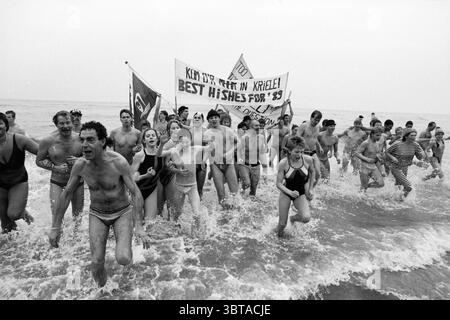 Silvester Tauchgang Zandvoort Neujahr Zandvoort, Whizgle News, Dutch Desk, Niederlande, 1950 - 2000 am 01-01-1989. Diese Themen werden in der Abbildung angezeigt. Eine lebhafte Szene entfaltet sich am Strand, wo eine große Gruppe von Menschen energisch auf die Wellen zuläuft. Die Individuen, unterschiedlich in Alter und Aussehen, plätschern durch das flache Wasser, ihre Ausdrucksformen sind eine Mischung aus Freude und Freude. Die meisten tragen Badeanzüge, die von einfarbigen Farben bis hin zu verspielten Mustern reichen, während einige von ihnen einzigartige Styles zeigen, wie z. B. ein gestreiftes Ensemble. Der Hintergrund zeigt einen gedämpften, bewölkten Himmel, der ein wenig so wirkt Stockfoto