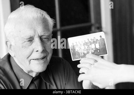 Herr H. Michel ehemaliger Fußballspieler, Whizgle News, Dutch Desk, Niederlande, 1950 - 2000, 08-07-1996. Diese Themen sind in der Abbildung dargestellt. Die Szene fängt einen warmen und nostalgischen Moment ein, den zwei Personen teilen. Ein älterer Mann mit einem Kopf aus weißem Haar und einem sanften Lächeln sitzt. Sein Ausdruck spiegelt ein Gefühl der Erinnerung wider, während er ein Foto vor sich sieht. Sein Gesicht zeigt Linien, die auf ein wohlgelebtes Leben hindeuten und Weisheit und Wärme ausstrahlen. Im Vordergrund greift eine Hand auf den Mann zu und präsentiert das Foto, das eine Gruppe von Menschen in einem historischen Bild zeigt Stockfoto