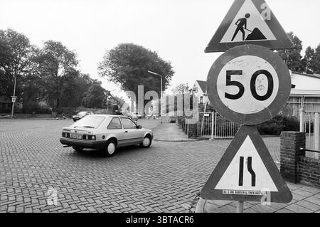 Schild „50 km“ Wijk aan Zeeërweg IJmuiden IJmuiden Wijk aan Zeeërweg Niederlande, Whizgle News, Dutch Desk, Niederlande, 1950 - 2000 am 07-09-1993. Dies sind die Themen im Bild. Die Szene fängt eine ruhige, urbane Kreuzung an einem bewölkten Tag ein, die von einer gedämpften Farbpalette geprägt ist, die von Grau- und Schwarztönen dominiert wird. Ein kleines Auto auf der linken Seite befindet sich in der Mitte der Kurve, dessen silberner Farbton sich dezent von der düsteren Umgebung abhebt. Die Linienführung des Fahrzeugs ist schlank und doch schlicht und zeigt einen nostalgischen Charme. Im Vordergrund befinden sich zwei große Warnzeichen. Der erste, indikati Stockfoto